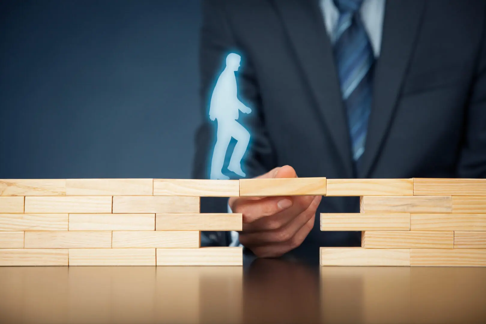 A person in a business suit holding up a block to form a bridge for a blue human silhouette to walk over